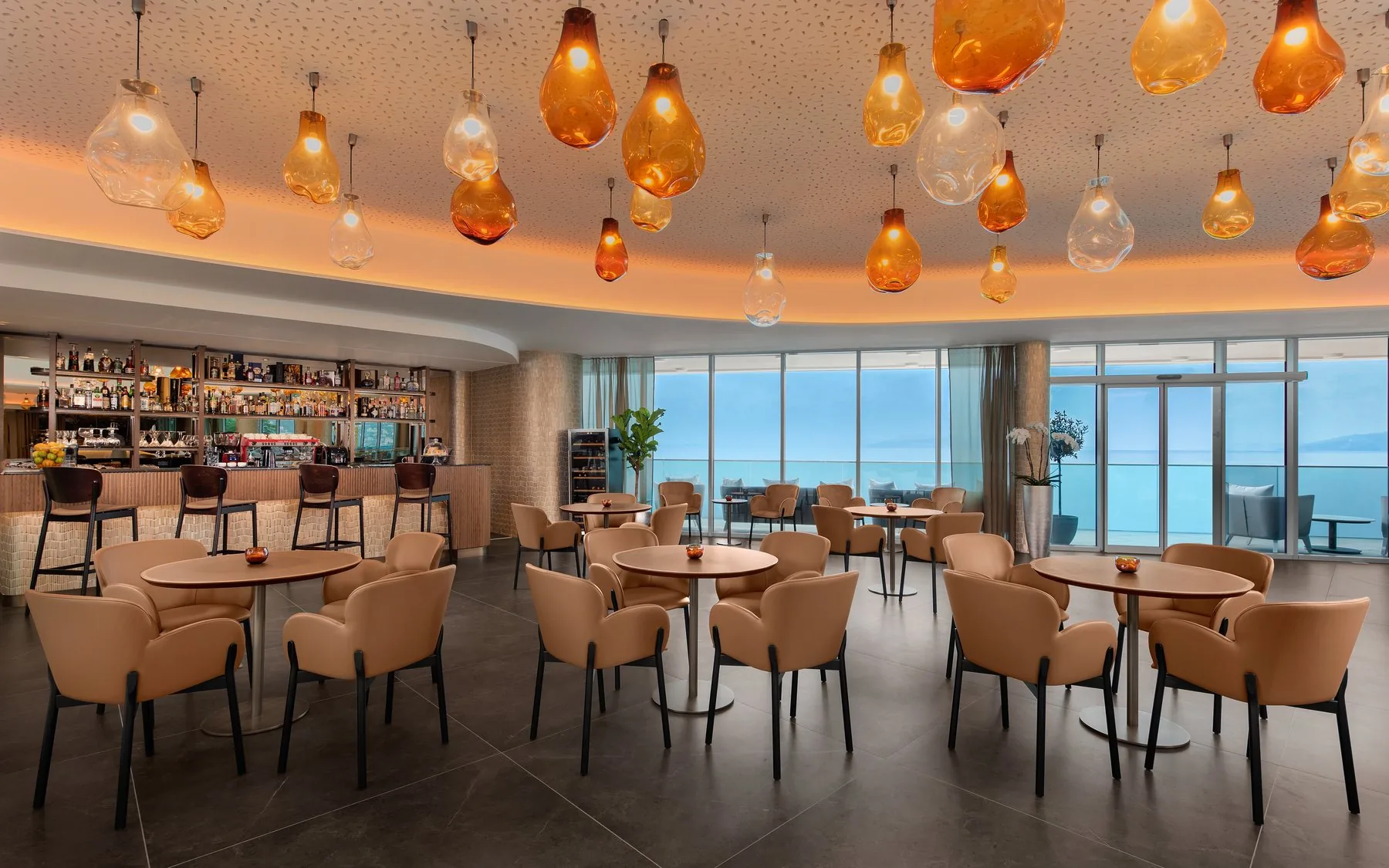 Sala panoramica di un hotel con vetrate sul mare, lampade in vetro soffiato e pavimento in pietra di grande formato