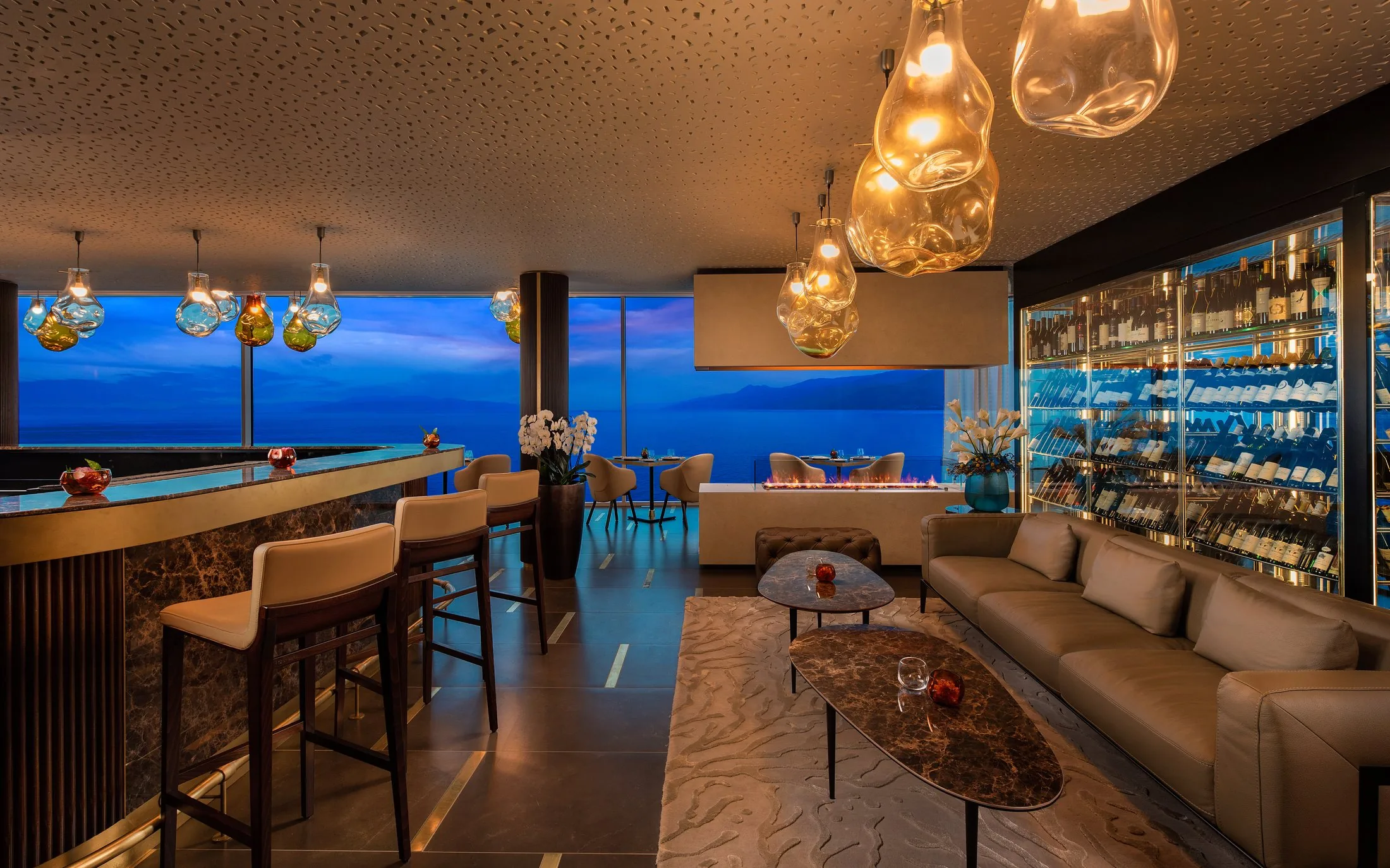 Lounge bar al tramonto affacciato sul mare: bancone rivestito in marmo scuro venato, profilo in ottone, tavolini in marmo