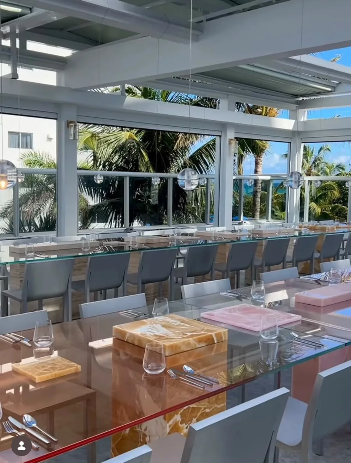 Sala di un ristorante affacciata sul mare: tavoli in vetro su basi in onice, con piatti in marmo rosa e miele