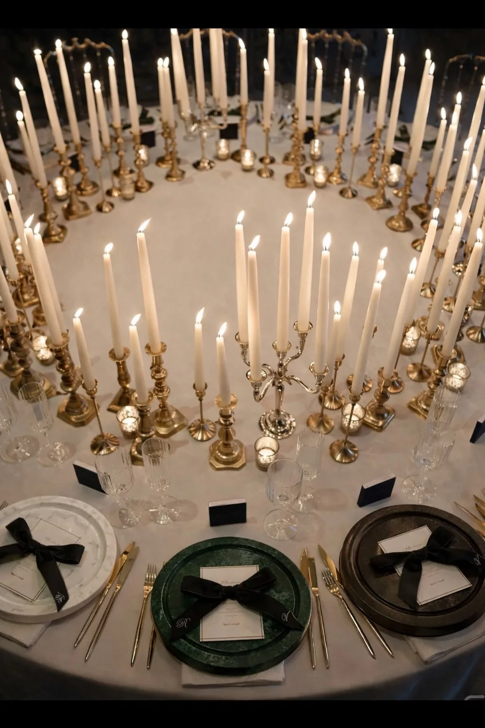 Tavola rotonda da banchetto illuminata a candele, con piatti in marmo bianco, verde alpi e bruno